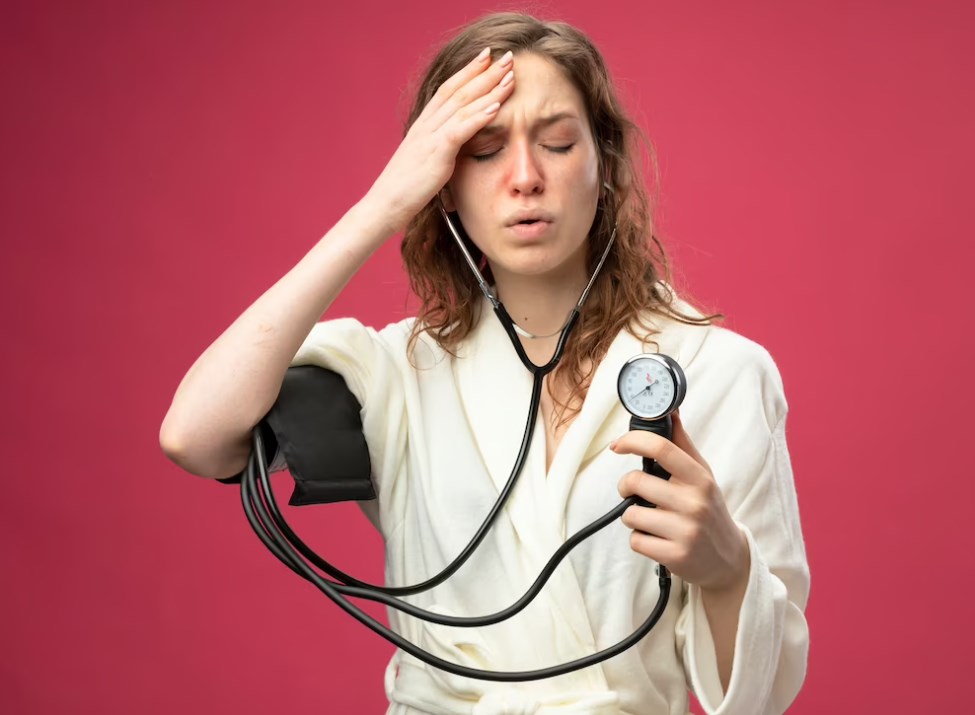 a weak young girl with closed eyes wearing a white robe measuring her own pressure with a sphygmomanometer while keeping her hand on her forehead