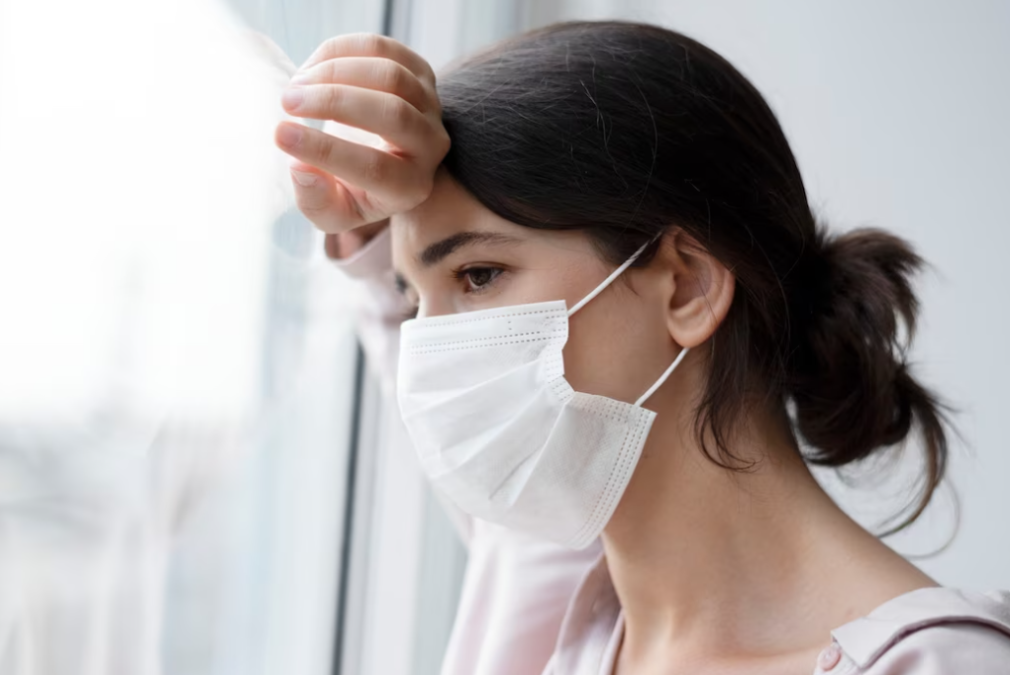 woman with hair tied in a bun in white mask leaning on the window and looking outside