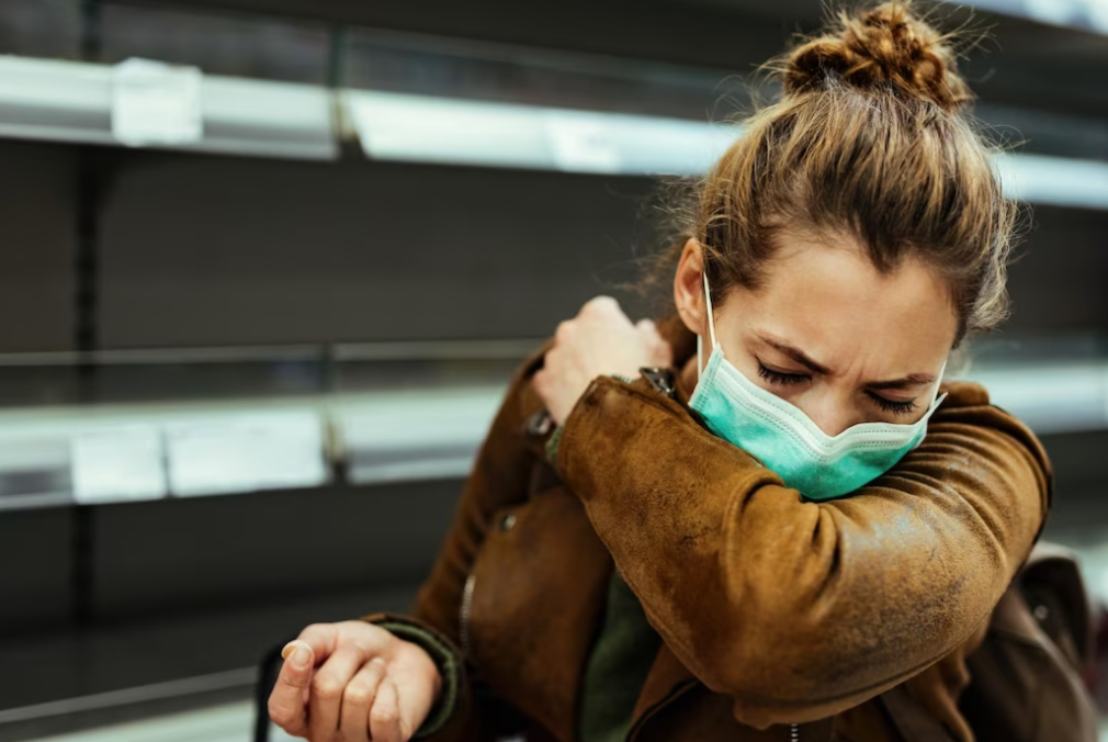 woman in a brown leather jacket and green mask sneezing into her hand